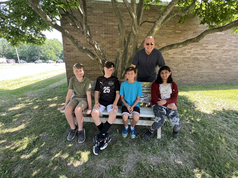 Four students sitting on bench outside with teacher standing behind